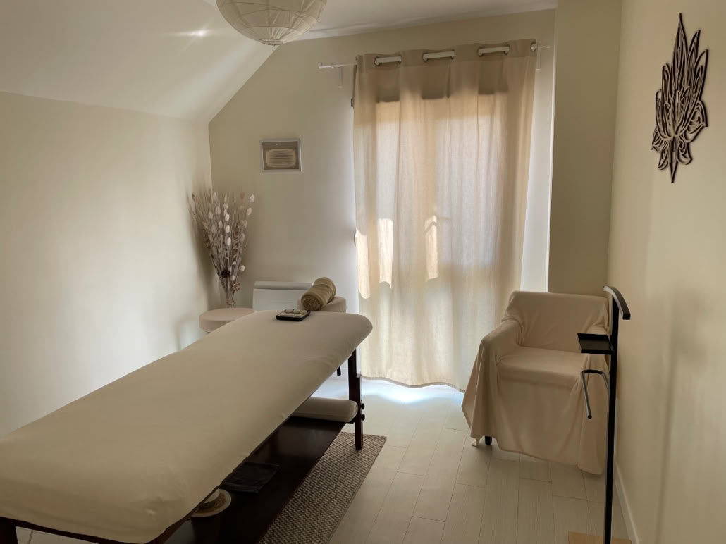 Salle de massage avec une table recouverte d'un drap blanc, une chaise beige, un rideau translucide à la fenêtre, et une décoration murale.